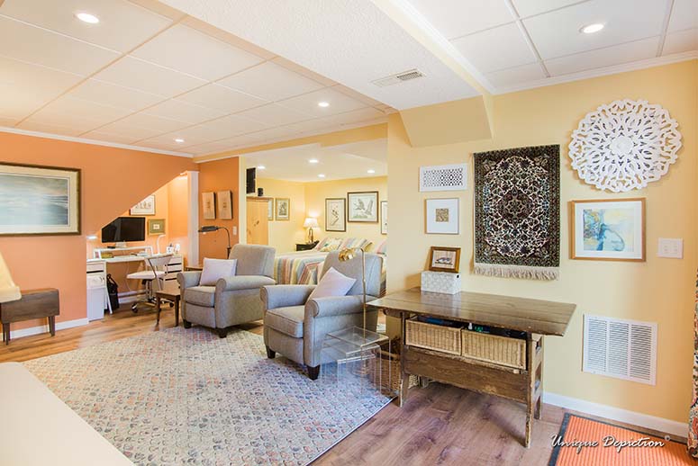 Finished basement living room with light yellow and light orange walls, area rug, cushioned chairs, side table, wall art.