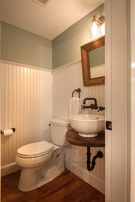 Powder room with vertical white-paneled wainscoting and vessel sink with rustic wooden base and black pipes/faucet