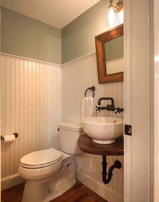 Powder room with vertical white-paneled wainscoting and vessel sink with rustic wooden base and black pipes/faucet