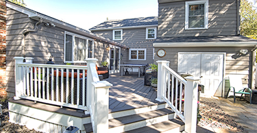 Back exterior view of a two-story gray home with raised wood deck with white railings and few steps in foreground leading to a patio area.