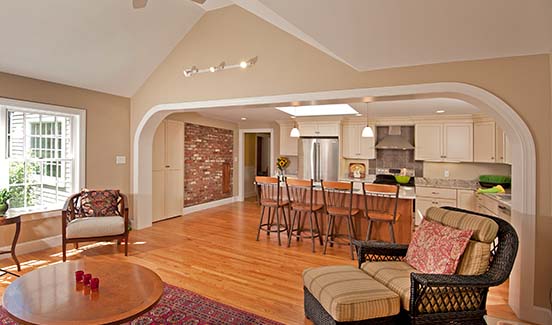 Renovated home addition with large open doorway from kitchen with island to living area. Wood floors. Light tan walls. Track lighting.