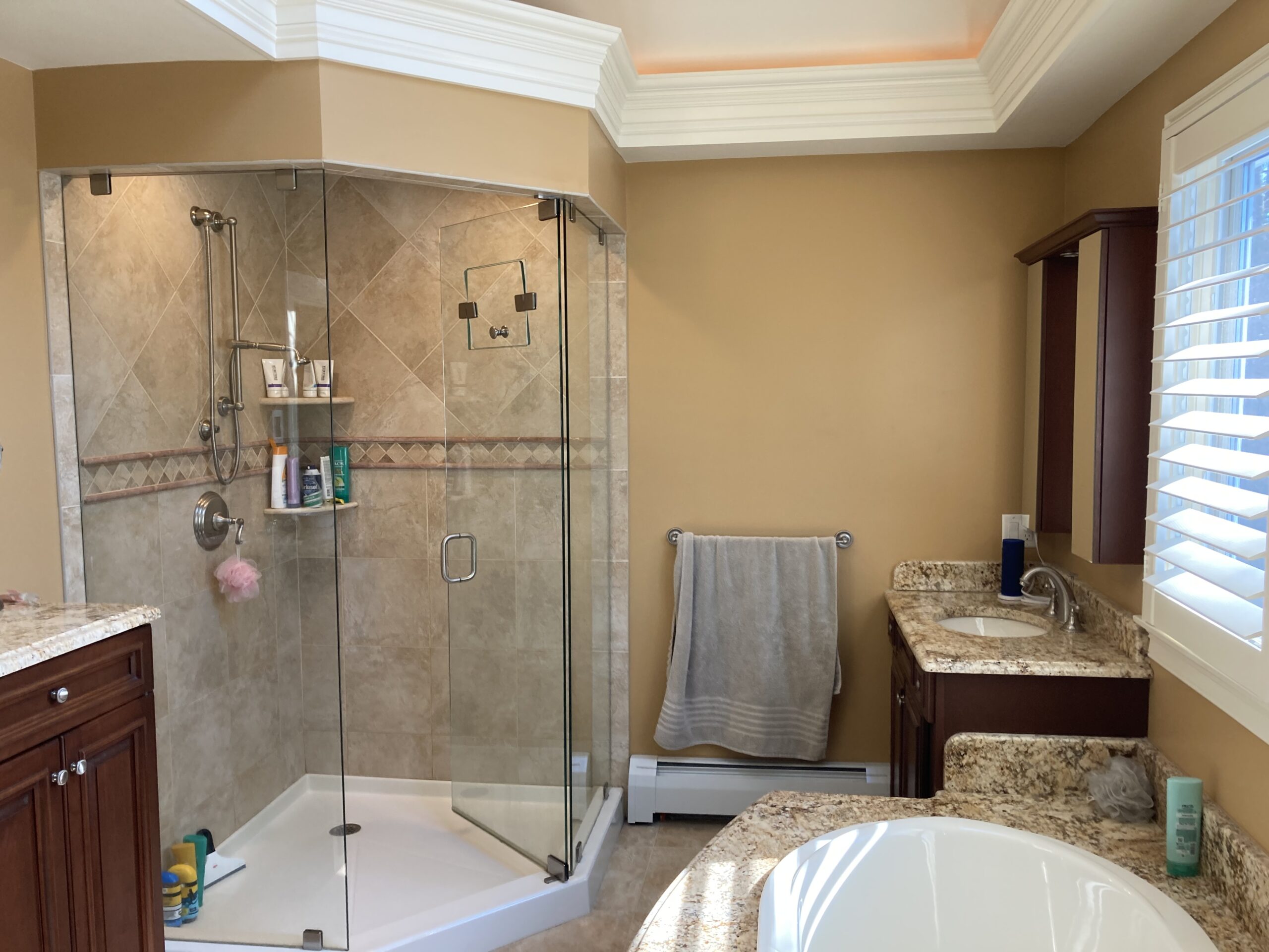 Tan-tiled standing shower with a glass door in a bathroom with granite sink countertops