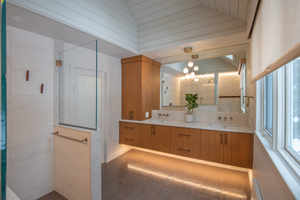 Large updated bathroom renovation with tan tile floors, woodgrain dual vanity with white stone countertops, and glass-enclosed shower area.