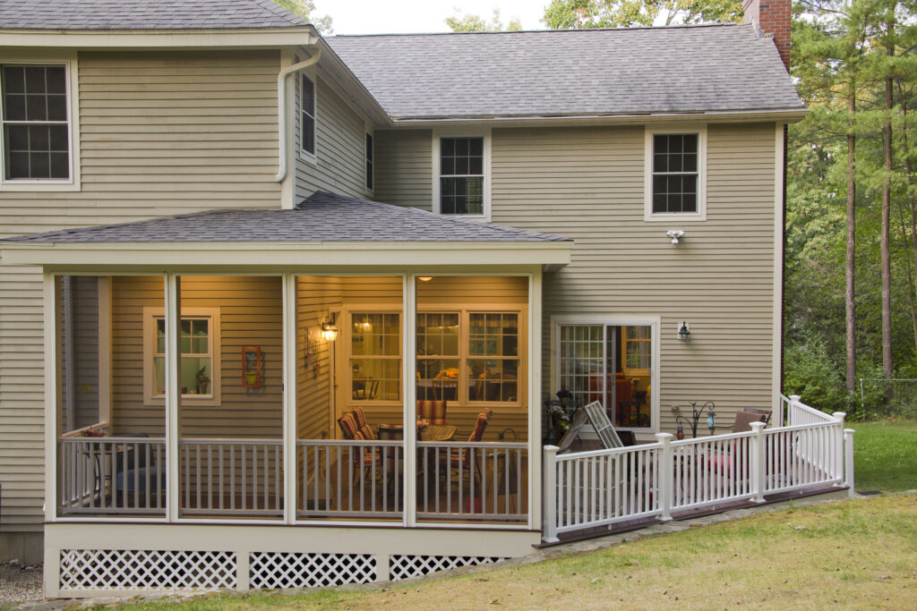 Screened-in porch added to the back of a large two-story beige home, with open deck to the right side, lights on in the porch, trees behind home.