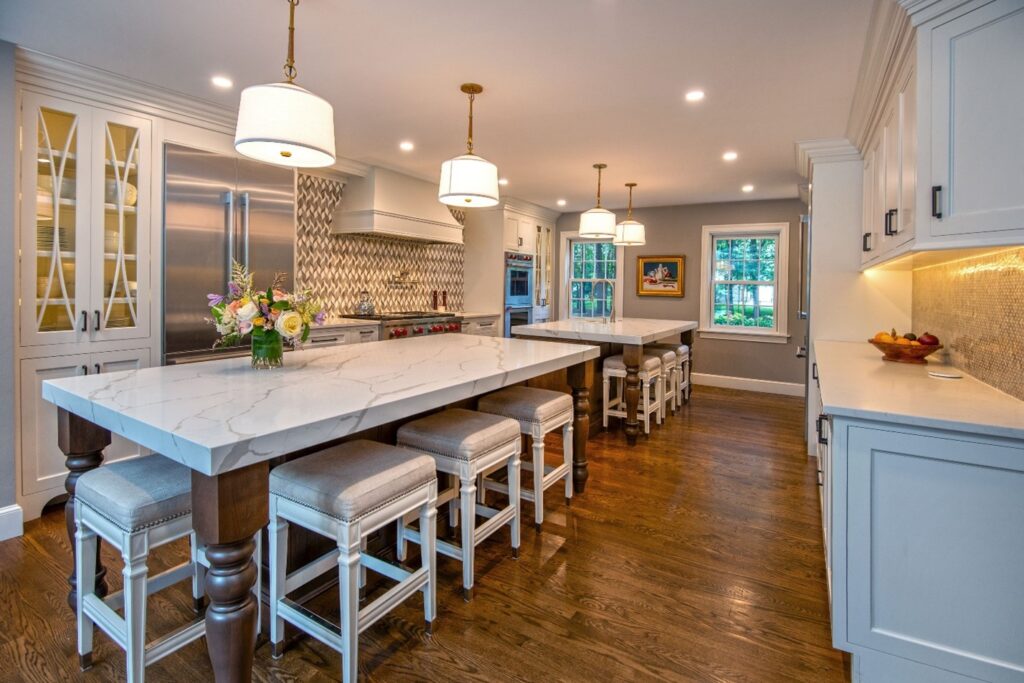 Remodeled kitchen with two long islands with stool seating, white marble slab countertops, wood floors, white cabinets along the wall, recessed lighting, window and side door on far wall.
