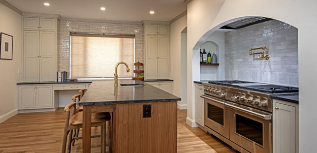 Remodeled kitchen with wood floors. woodgrain island and white cabinets against far wall, black soapstone countertop, double range/ovens along right side with white brick backsplash.